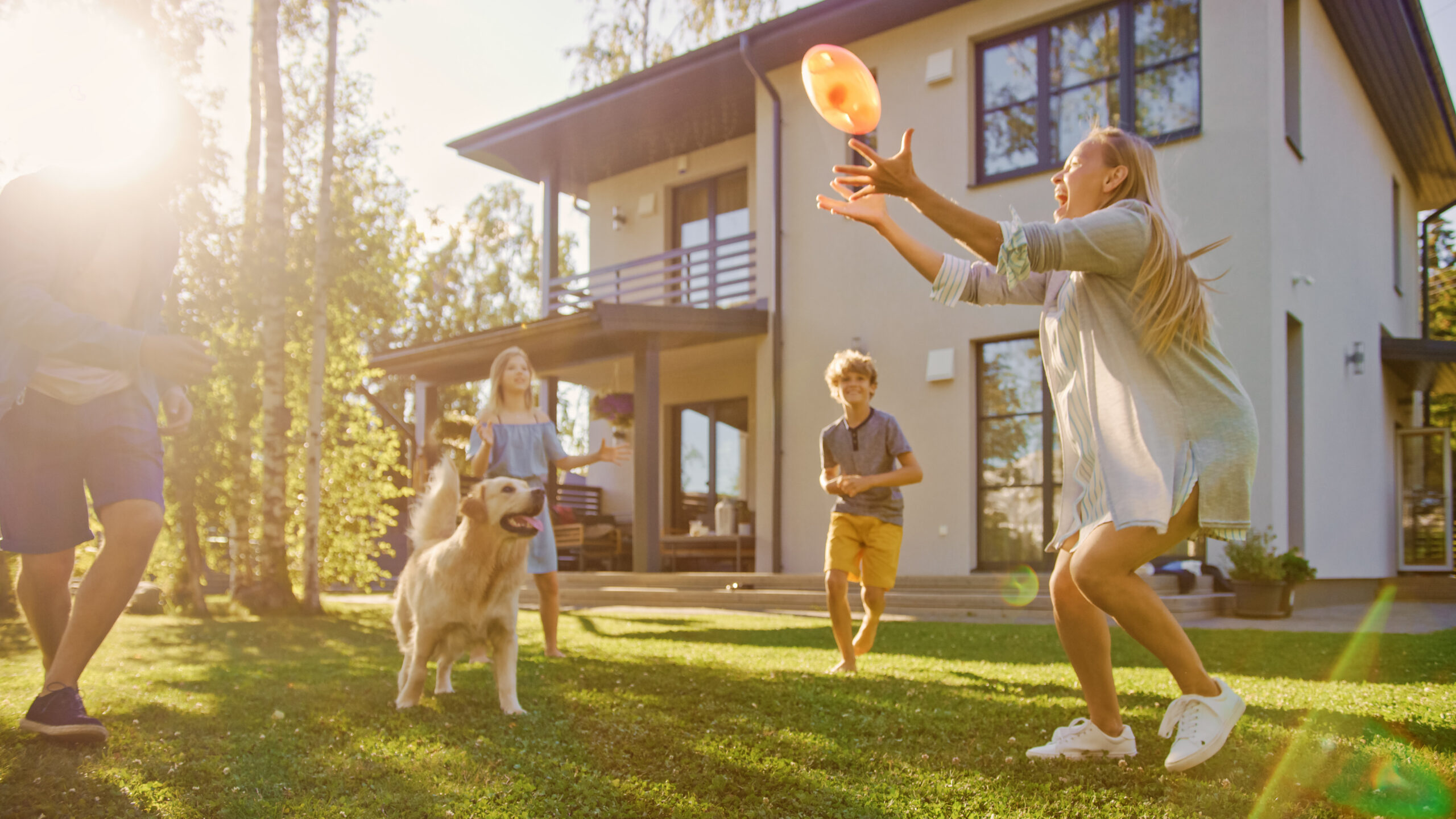 Eine Familie spielt mit einem Ball auf einer Rasenfläche vor einem modernen Einfamilienhaus. Ein Hund befindet sich ebenfalls im Garten.